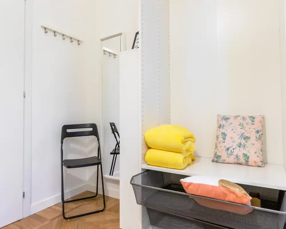 Entrada luminosa y ordenada con una silla plegable, perchas de pared, un espejo de cuerpo entero y estanterías organizadas con toallas dobladas y cojines — ideal para mostrar una entrada acogedora.