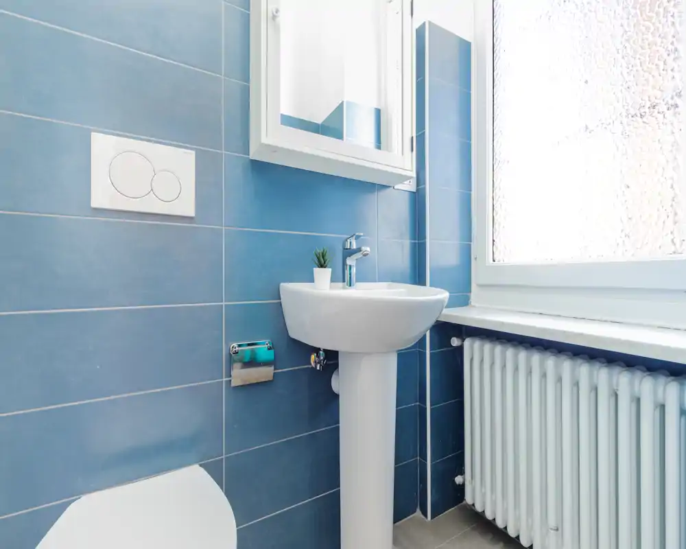 Baño limpio y luminoso con lavabo de pedestal, pulsador de cisterna empotrado y ventana esmerilada que aporta luz natural; azulejos azules que transmiten modernidad y frescura.
