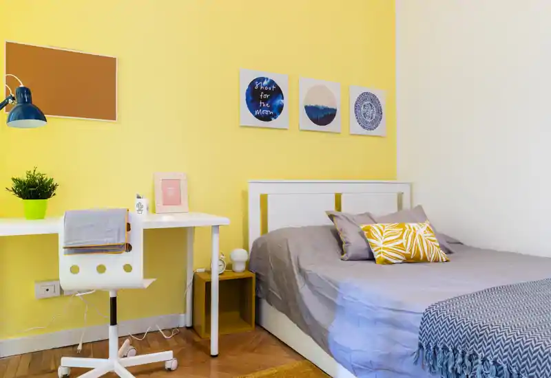 Habitación individual moderna y luminosa con cabecero blanco, ropa de cama gris y cojines decorativos amarillos. Un escritorio blanco compacto y una silla junto a la cama, frente a una pared amarilla alegre, crean un espacio brillante y ordenado, ideal para estudiantes o alquileres temporales.