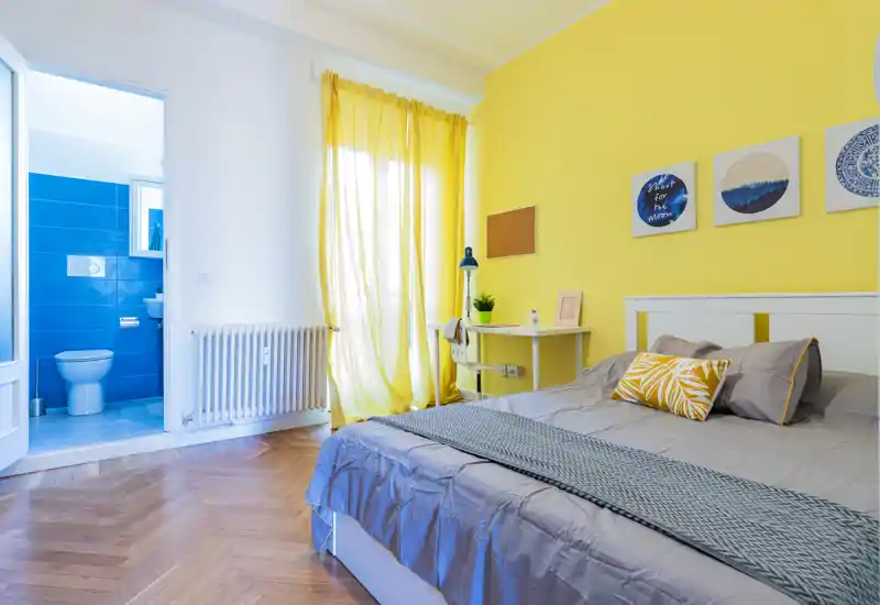 Dormitorio moderno y luminoso con cama doble, pared amarilla cálida y un pequeño escritorio; baño en suite con azulejos azules visible por la puerta abierta.