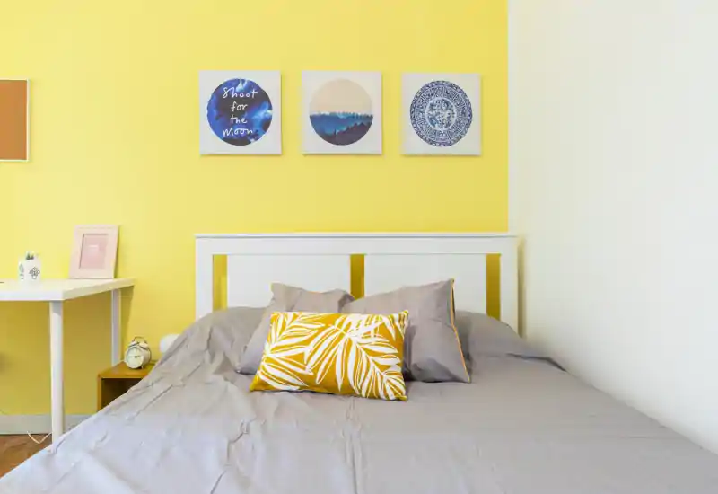 Dormitorio moderno y luminoso con cabecero blanco, ropa de cama gris y un cojín amarillo decorativo. Tres cuadros sobre la cama y una pequeña mesa lateral crean un ambiente alegre y bien iluminado.