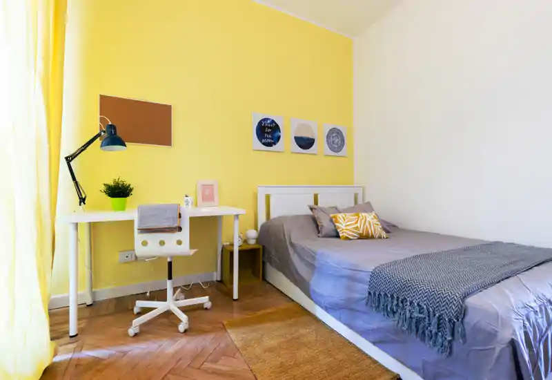 Dormitorio individual luminoso y bien presentado con cama cómoda, cojines decorativos y un escritorio ordenado junto a la ventana; pared amarilla y parquet aportan una atmósfera acogedora.