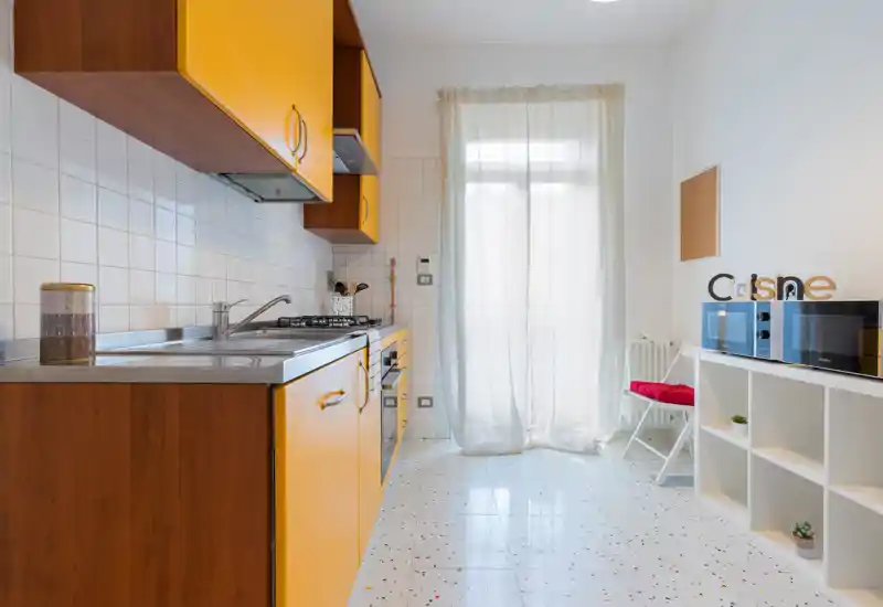 Bright, compact kitchen with yellow cabinetry, stainless sink and gas hob; natural light from a sheer-covered door makes the space inviting and functional.