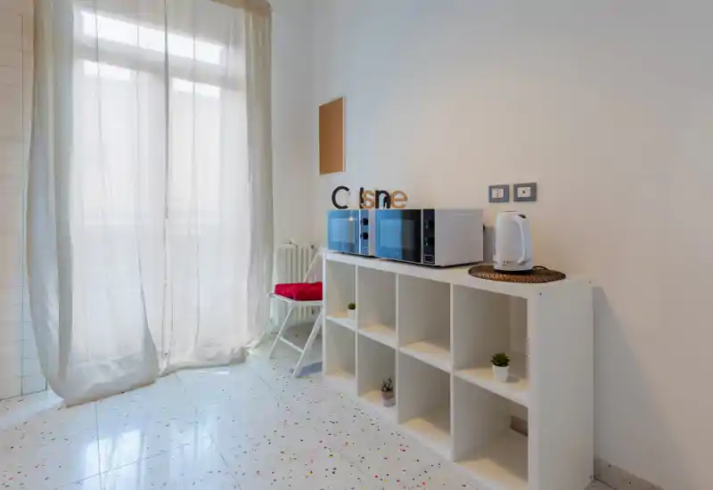 Compact, bright kitchenette with microwave and electric kettle on a white shelving unit; sheer curtains diffuse natural light and small decorative plants add a welcoming touch.