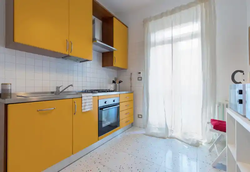Bright, compact kitchen with bold yellow cabinetry, stainless sink, gas hob and an oven. Large sheer-covered window/door floods the space with natural light, and terrazzo-style floor adds character.
