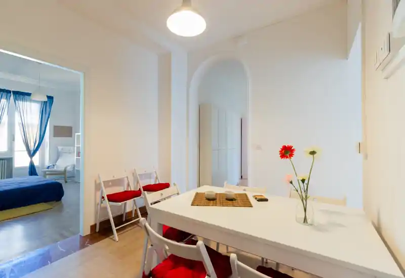 Bright, modern dining corner of an open living area with a white table, folding chairs with red cushions and decorative flowers; the bedroom is visible through a wide doorway.