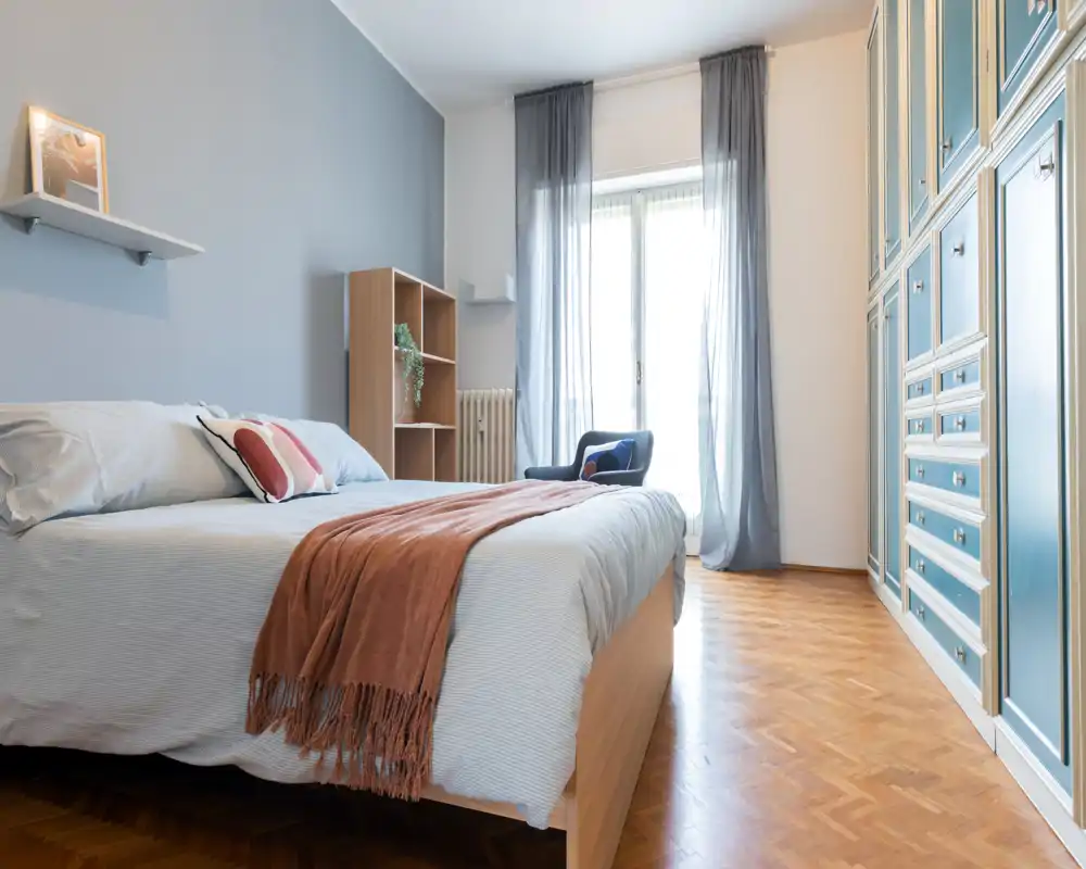 Camera da letto luminosa e di stile con un ampio letto, biancheria morbida, plaid caldo e spaziosi armadi a muro. La luce naturale dalla finestra rende l'ambiente arioso e accogliente, perfetto per annunci.