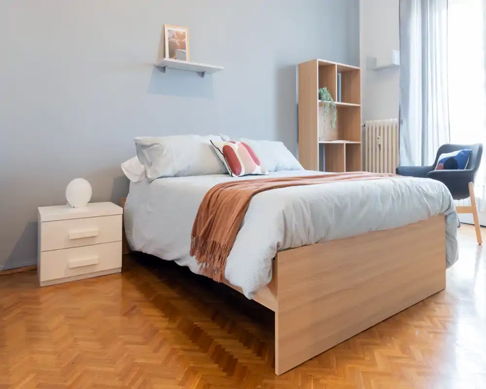 Camera da letto luminosa e moderna con ampio letto in legno, biancheria morbida, cuscini decorativi e un plaid accogliente. La luce naturale illumina la stanza; mensole semplici e una poltrona da lettura completano l'arredo minimal.