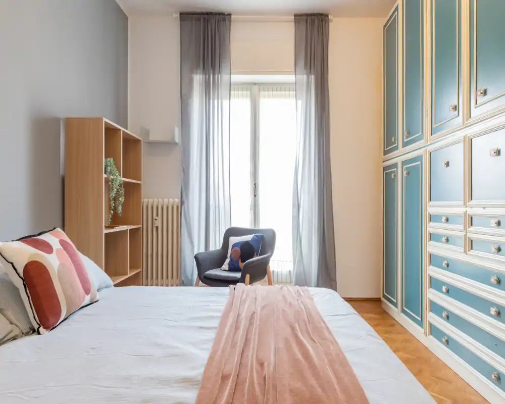 Camera da letto luminosa e curata con un ampio letto in primo piano, cuscini decorativi e un copriletto morbido; armadi a muro blu e luce naturale che illumina la stanza.