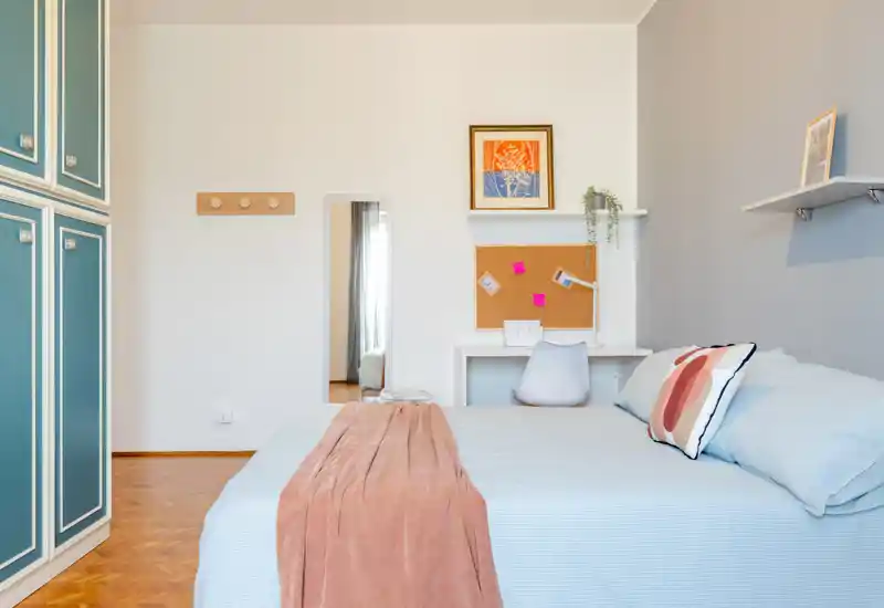 Bright, cozy bedroom with a double bed dressed in pale linens, decorative cushions and a warm throw; built-in wardrobe and a small desk create a functional, stylish space.