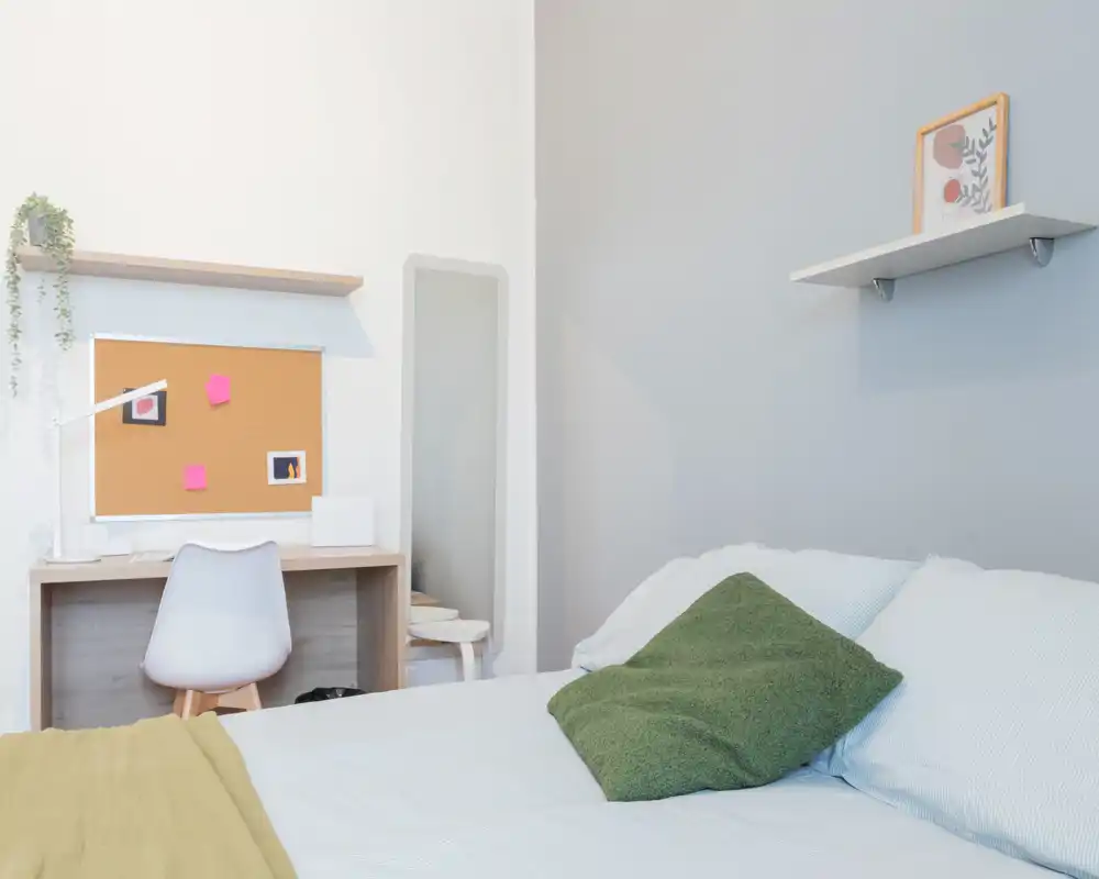 Camera da letto luminosa e minimal con letto in lenzuola chiare, cuscino verde come dettaglio e una piccola scrivania con bacheca: ideale per un affitto funzionale e accogliente.