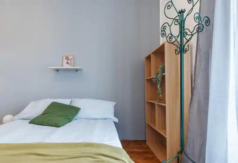 Minimal, well-lit bedroom with a double bed dressed in white linens, a green accent cushion and a simple shelf above the headboard; wooden shelving and parquet floor add warmth.