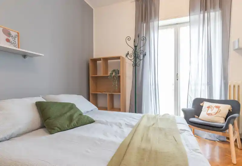 Bright, cozy bedroom with a large bed, soft linens, a comfy armchair and natural light from a balcony door.