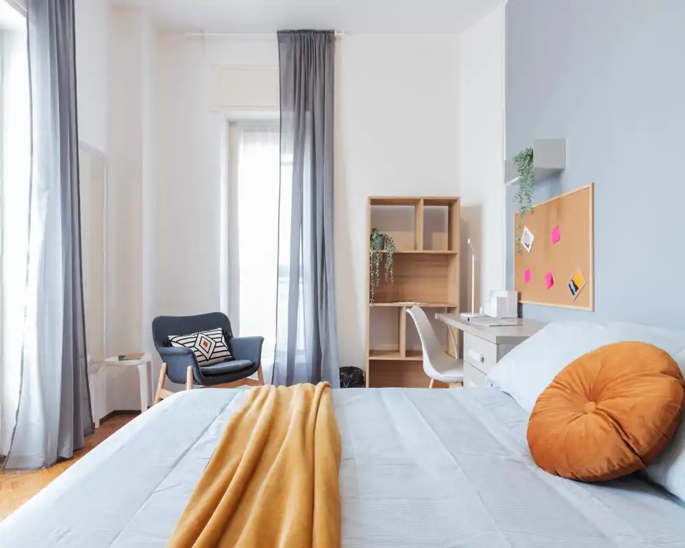 Bright, modern bedroom with a large bed in the foreground, warm mustard throw and round cushion, a cozy armchair by a tall window and a small desk with shelving—ideal for a comfortable rental listing.