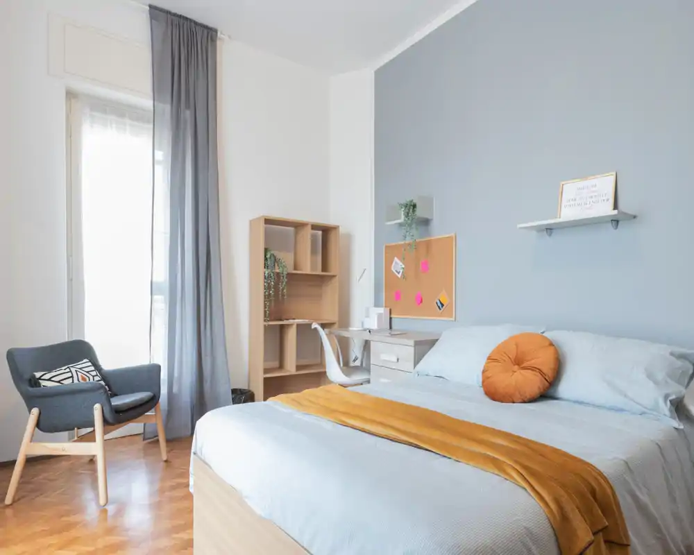 Bright, well-styled bedroom with a double bed dressed in light linens, a warm mustard throw and a decorative round cushion; workspace and shelving beside the bed and a cozy armchair by the window.