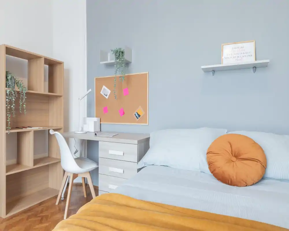 Bright, modern bedroom with a neatly made bed, soft blue linens, a mustard throw and a round decorative cushion; a compact desk and shelving create a practical study corner.