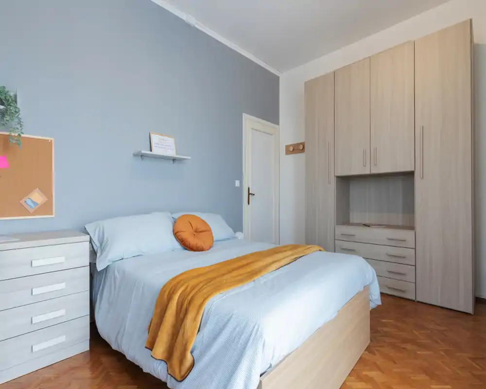 Bright, well-styled bedroom with a double bed dressed in light linens, a warm ochre throw and matching round cushion, plus fitted wardrobe and bedside drawers.