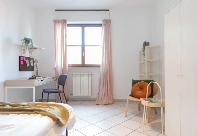 Chambre lumineuse au style doux avec lit visible au premier plan, bureau près de la fenêtre et coussins chauds apportant une ambiance cosy et minimaliste.