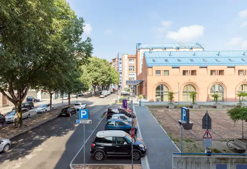 Vue de rue ensoleillée avec stationnement sur voirie et un bâtiment en brique au toit bleu ; lumière du jour claire et arbres ombrageant la route, idéale pour présenter le quartier et ses commodités.