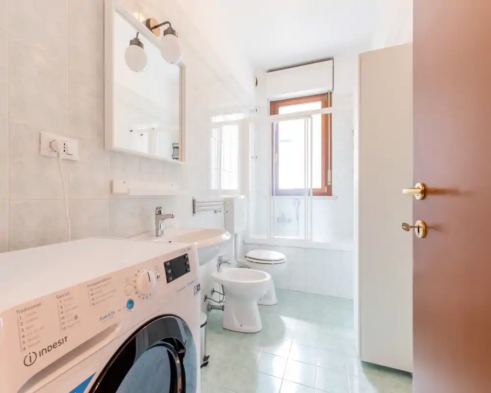 Salle de bains lumineuse et soignée avec lavabo, bidet, toilettes et baignoire avec paroi vitrée ; une machine à laver est placée près du lavabo et une grande fenêtre apporte la lumière naturelle.