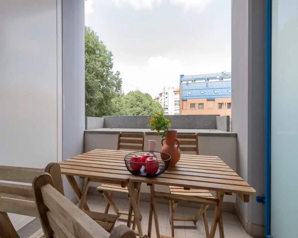 Balcon ensoleillé avec table et chaises pliantes en bois, décoré d'un vase et d'un panier de pommes — parfait pour un repas en plein air ou un café.