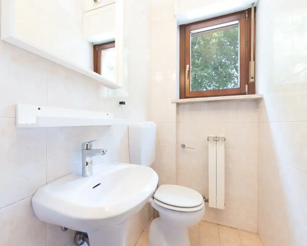 Salle de bains compacte et lumineuse avec lavabo blanc, WC et une fenêtre à cadre en bois apportant la lumière naturelle.