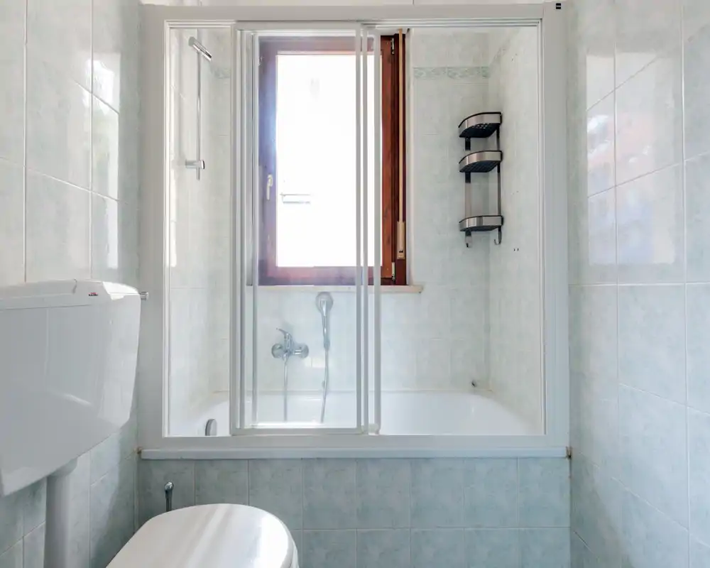 Salle de bains compacte et lumineuse avec baignoire et paroi de douche coulissante en verre, carrelage mural et fenêtre apportant la lumière naturelle.