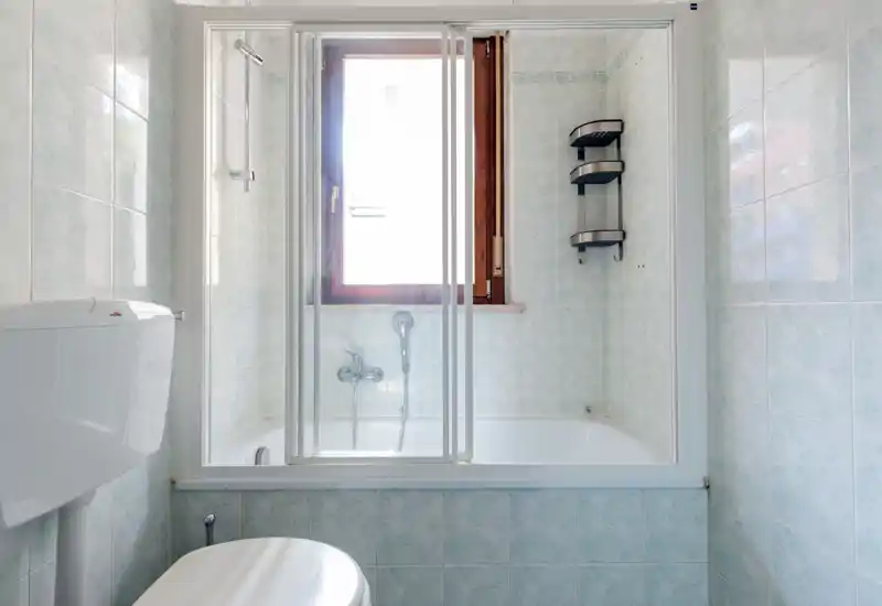 Small tiled bathroom with a bathtub and sliding shower screen, wall-mounted toilet cistern, and a window providing natural light.