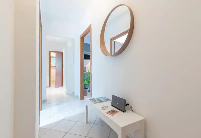 Bright, minimalist entrance hallway with a white bench, decorative round mirror and tiled floor — welcoming and well-lit arrival space.