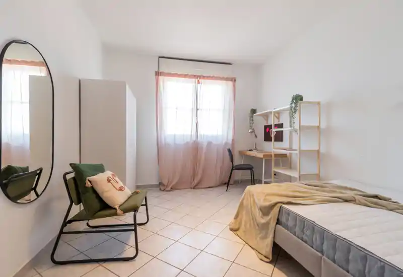 Bright, minimal bedroom with a single bed, a reading chair, a desk and shelving. Soft curtains and neutral tones create a calm, airy atmosphere ideal for a student or solo renter.