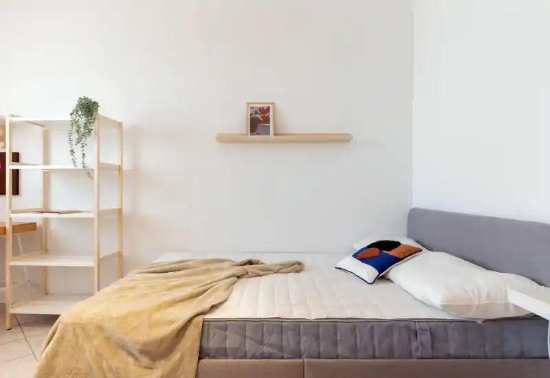 Bright, minimal bedroom with a neatly made double bed, soft throw and a decorative cushion; simple shelving adds functional storage and a calm, airy feel.