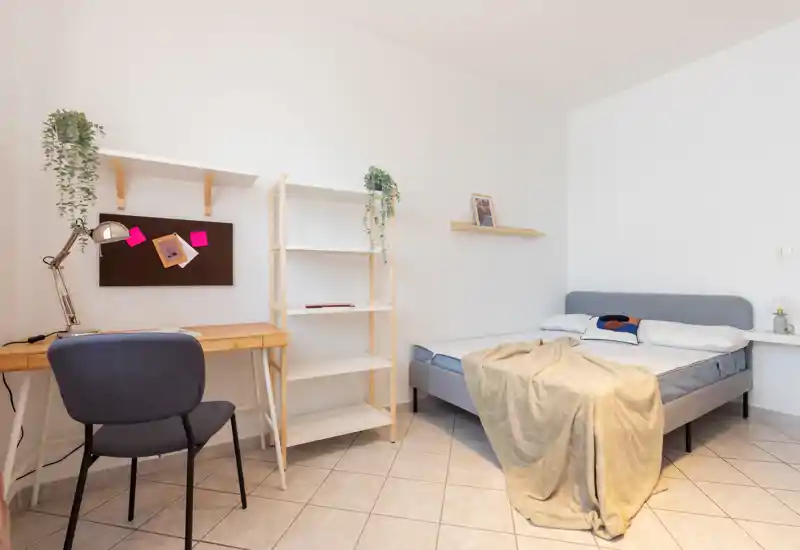 Bright, minimalist bedroom with a double bed, soft throw and two pillows; a small desk and shelving create a functional work corner.