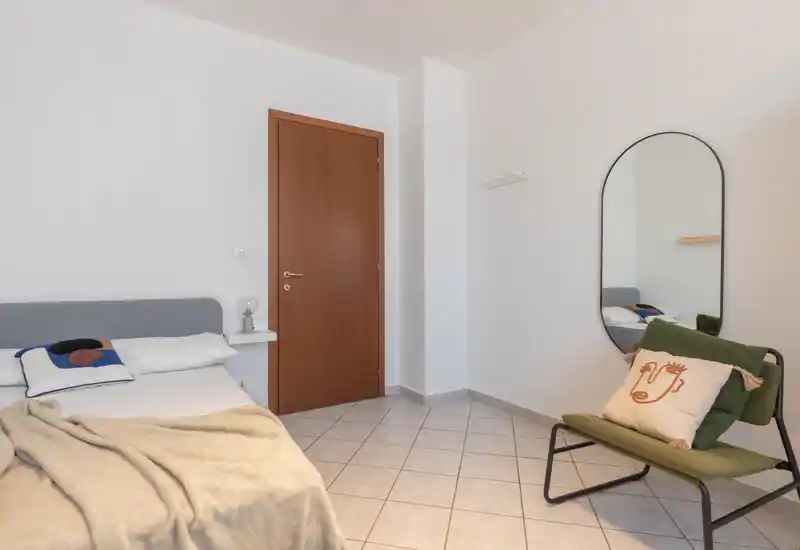 Bright, minimal bedroom with a partially visible double bed, neutral linens and a cozy throw, plus a mirror and an accent chair.