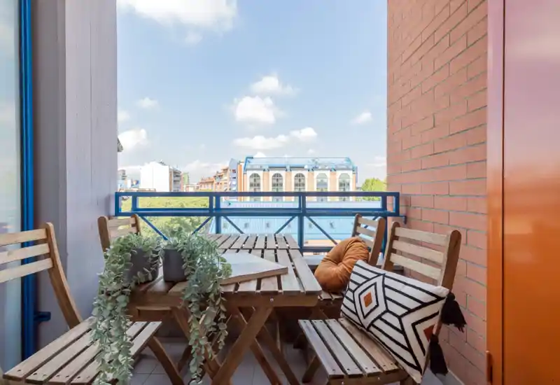 Sunny furnished balcony with a wooden table and chairs, decorative cushions and potted plants, overlooking nearby buildings — ideal for outdoor dining or relaxing.