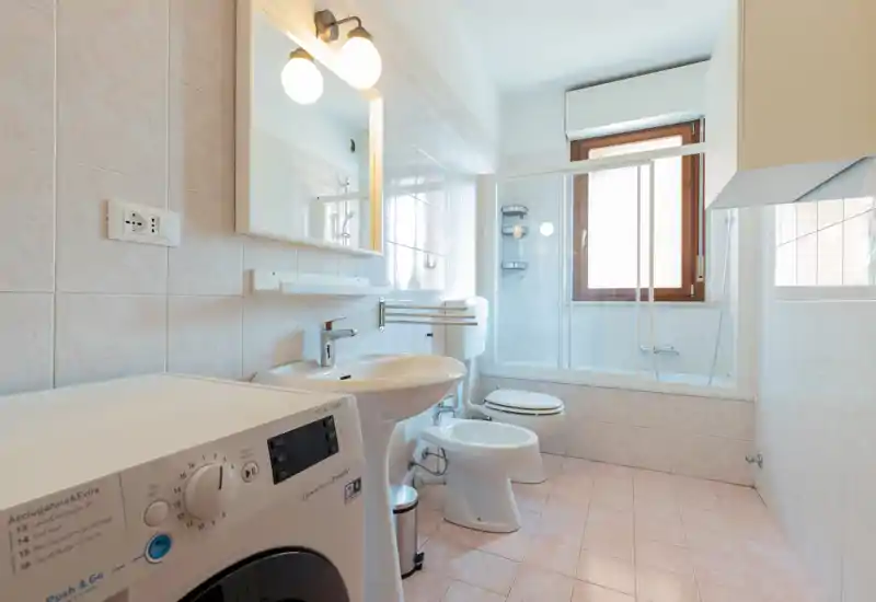 Bright, clean bathroom with a sink, bidet, toilet and a combined bathtub/shower behind glass doors; a washing machine is visible in the foreground and a window provides natural light.