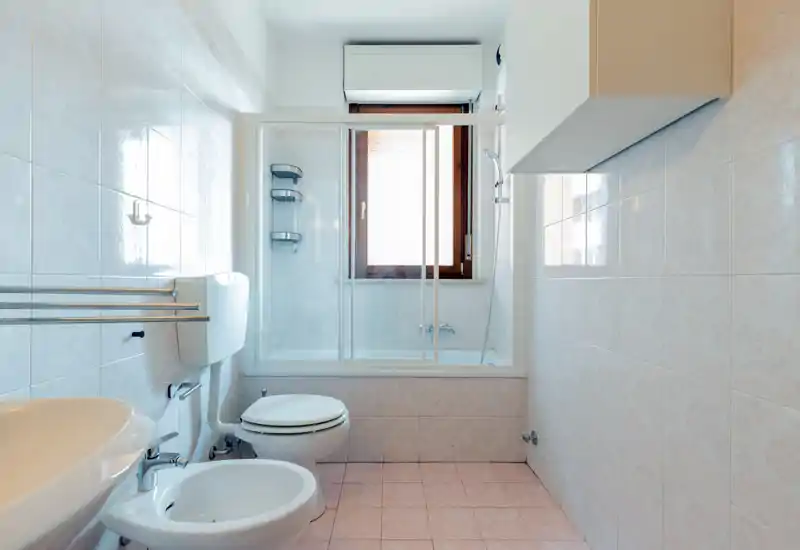 Bright, clean bathroom with sink, bidet, toilet and bathtub with sliding glass doors; tiled walls and floor and a window providing natural light.
