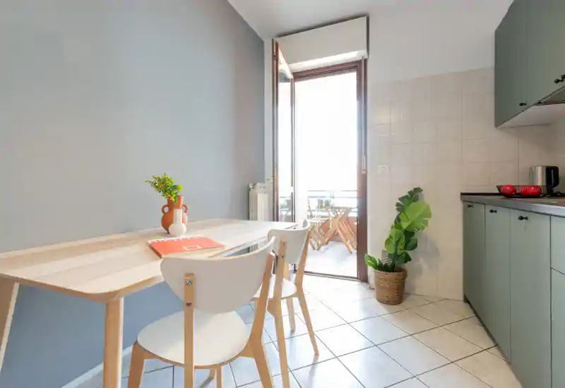 Bright modern kitchen with a small dining table by the balcony. Mint-green cabinets, a wooden table with two chairs and decorative plants create a fresh, welcoming space with plenty of natural light.