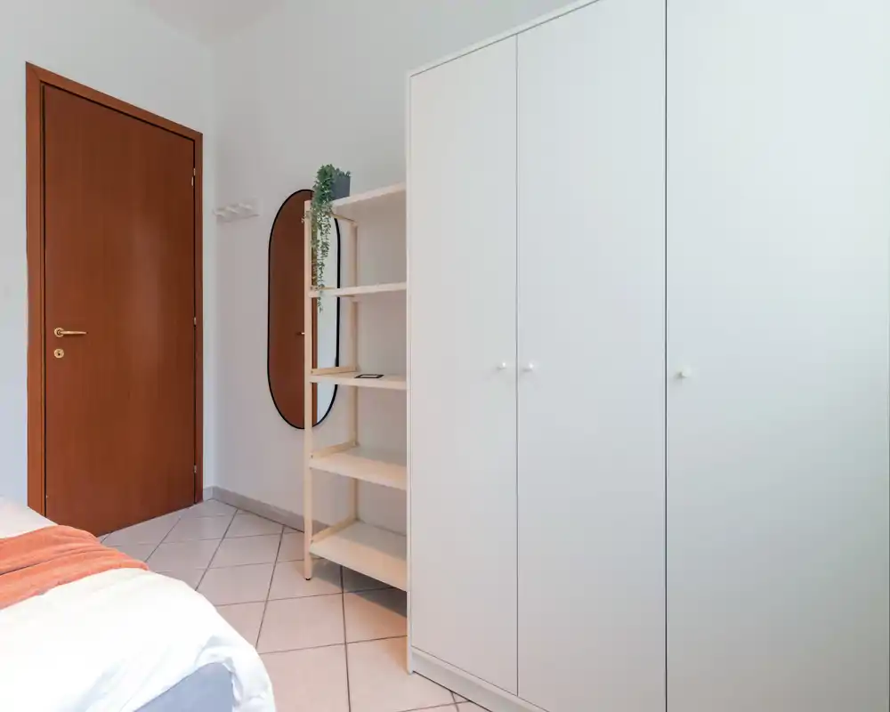 Coin de chambre minimaliste et lumineux avec le bord du lit visible, une grande armoire blanche, des étagères ouvertes et un miroir mural — propre et moderne.