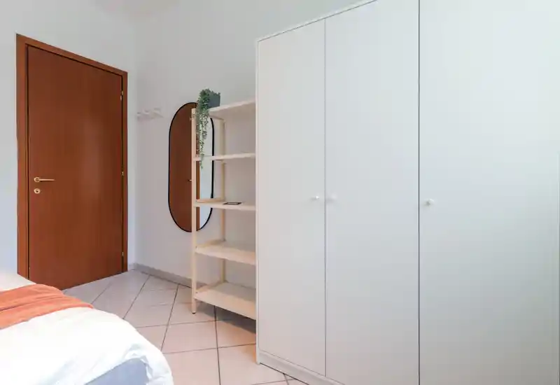 Coin de chambre minimaliste et lumineux avec le bord du lit visible, une grande armoire blanche, des étagères ouvertes et un miroir mural — propre et moderne.