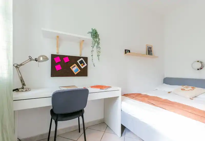 Chambre lumineuse et minimaliste avec un lit simple, un bureau soigné et une lumière naturelle — idéale pour un étudiant ou une personne seule. Murs blancs, textiles doux et un espace de travail simple créent une atmosphère calme et fonctionnelle.