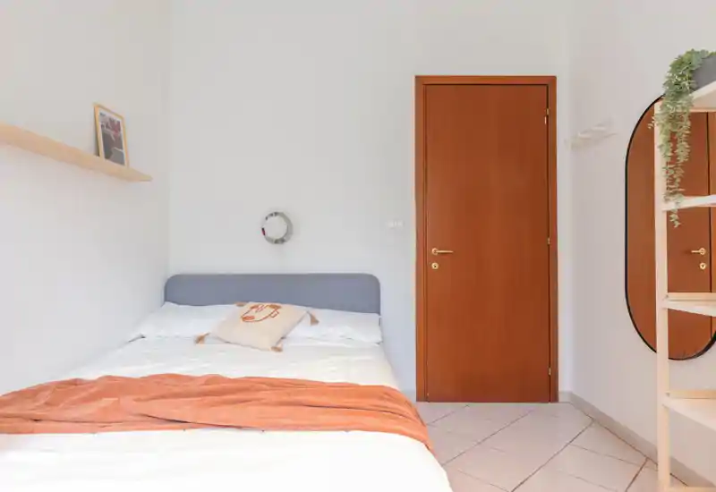 Chambre lumineuse et minimaliste avec lit bien fait, plaid terracotta et coussin décoratif. La pièce dispose d'une porte en bois, d'un miroir en pied et d'une ambiance claire et accueillante.