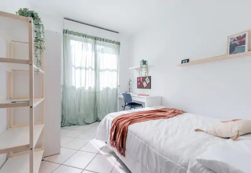 Chambre lumineuse et minimaliste avec un lit simple bien fait, un bureau près de la fenêtre et une douce lumière naturelle filtrant à travers les voilages.