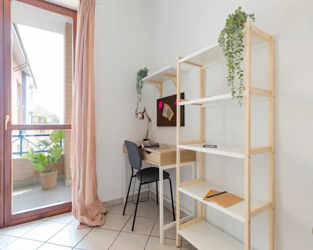 Bright home office corner with a small wooden desk, chair, open shelving and a balcony door that brings abundant natural light—simple, airy and practical for remote work.