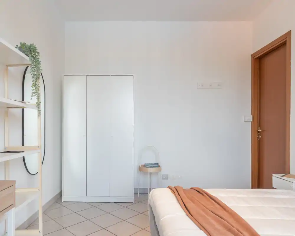 Bright, minimalist bedroom with a neatly made bed in the foreground, a white wardrobe and simple shelving. The room feels airy and clean with warm wooden accents and soft textiles.