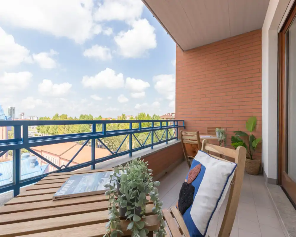 Sunny furnished balcony with wooden seating, potted plants and a distant treetop view — ideal for relaxing outdoors.
