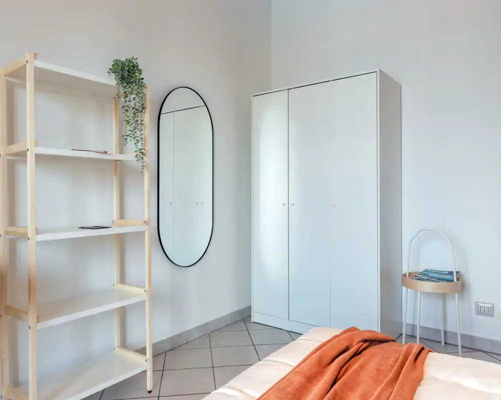 Bright, minimal bedroom with a visible bed corner, white wardrobe, wall mirror and shelving—clean styling and warm throw for a welcoming feel.