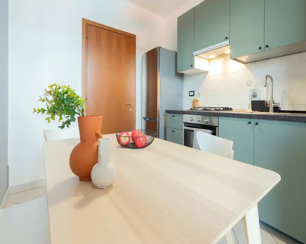 Bright, modern kitchen with muted green cabinets, a built-in oven and a dining table styled with vases and a fruit bowl—an inviting space for casual meals.