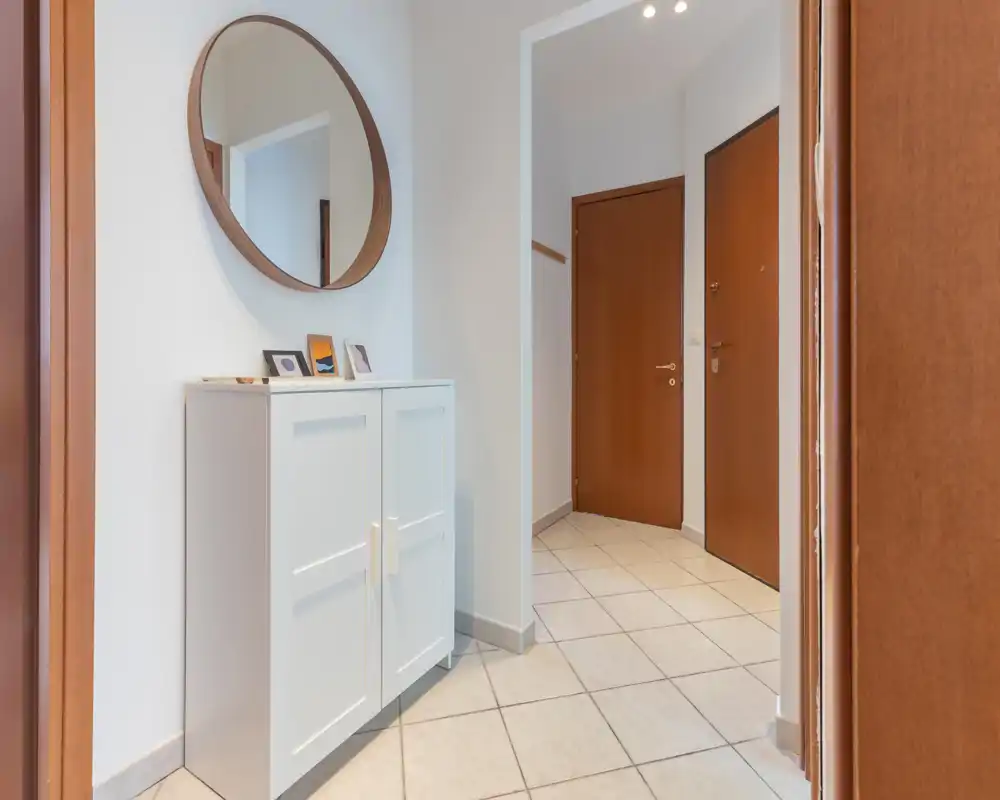 Bright, well-lit entrance with tiled floor, a white storage cabinet and a round wall mirror — neat and welcoming entryway ideal for first impressions.