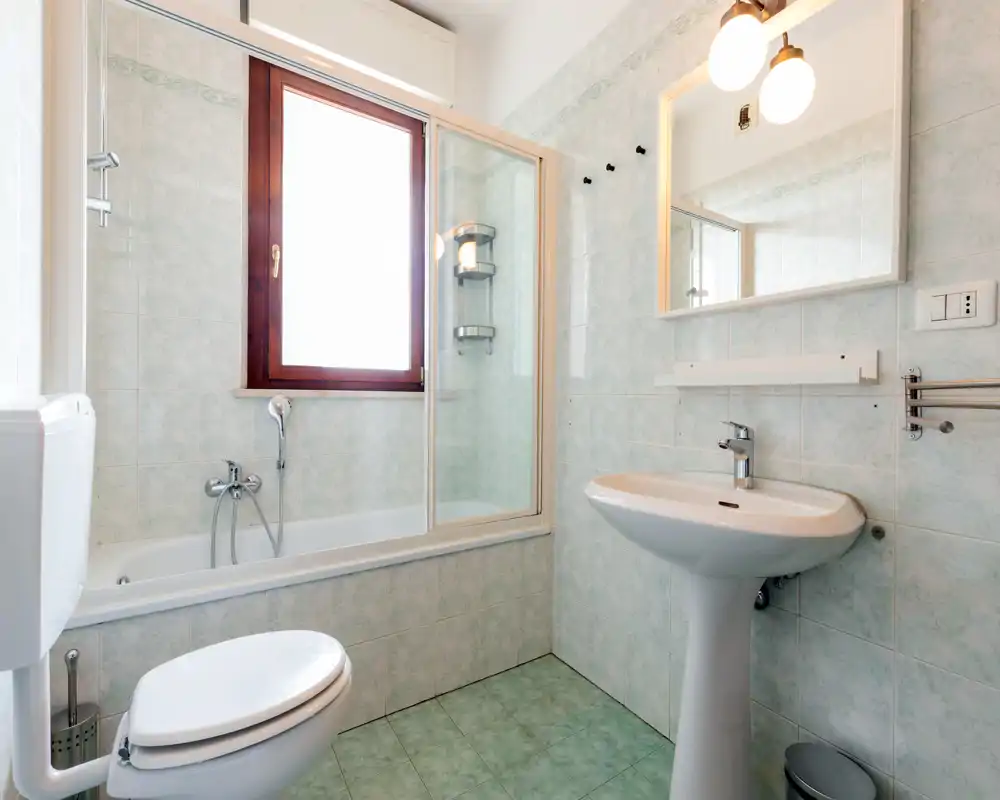 Bright, tiled bathroom with a bathtub and glass shower screen, pedestal sink and toilet. Natural light from a window makes the space feel clean and airy.