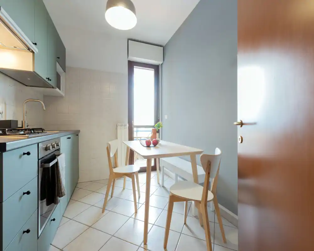 Bright compact kitchen with pastel cabinets, gas stove, and a small dining table. Natural light from the door and minimal, modern furnishings create a fresh, welcoming space ideal for a couple or solo occupant.
